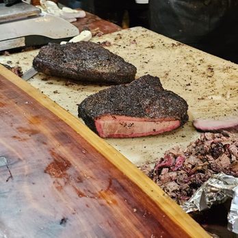 Mad Jack's Mountaintop Barbecue - Look at that smoke ring. - Two smoked brisket pieces with a dark bark are placed on a surface, one sliced to reveal a pink smoke ring and tender interior, with a meat slicer and gloves visible in the background. - Barbeque Near Me - Cloudcroft, New Mexico