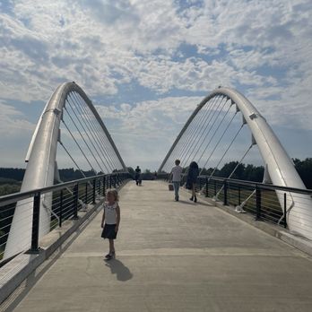 Riverfront Park - A concrete pedestrian bridge features large, white arch supports on either side, connected by cables. Several people, including a young girl in the foreground and adults further along, walk across the bridge. The sky above is partly cloudy, and the surrounding landscape includes trees and open sky, indicating an outdoor setting. - Parks Near Me - Salem, Oregon
