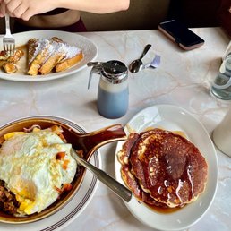 #4. Mexican Skillet I think - A breakfast meal includes a plate of pancakes with syrup, a skillet dish topped with a fried egg and vegetables, and a cup of black coffee. The table also has a water spray bottle, a spoon, a smartphone, and a small container of syrup. The setting appears to be in a restaurant with a marble tabletop and a window nearby.
