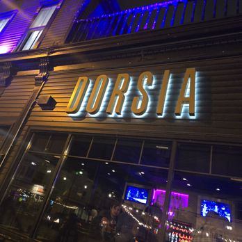 Dorsia - Restaurant Entrance - The image shows the exterior of Dorsia, a restaurant, at night. The building features a large illuminated sign spelling 'DORSIA' on its facade. The exterior has decorative architectural elements and is lit with colorful lights, including blue and purple accents. Through the large window, the interior is visible, with reflections of neon lights and some patrons inside. The scene captures a lively nighttime atmosphere with vibrant lighting. - Italian Near Me - Milwaukee, Wisconsin