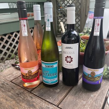 Table Wine - Four bottles of wine and sparkling wine are placed on a weathered wooden table outdoors. The bottles include rosé, white, and sparkling varieties, each with distinct labels and caps. The background features a garden area with a lattice fence, a deck, and some greenery, indicating an outdoor setting suitable for wine tasting or social gathering. - Beer, Wine & Spirits Near Me - Madison, Wisconsin