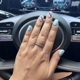 A hand with a diamond ring and light blue nail polish is placed on a car steering wheel, with a digital dashboard visible in the background.