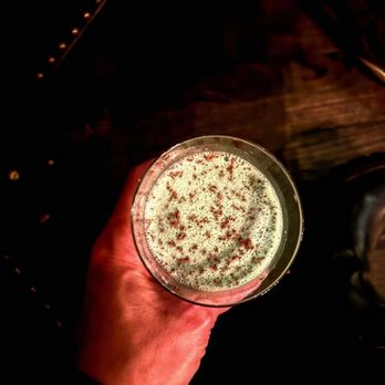 Highball - A person holds a cocktail glass filled with a frothy beverage, garnished with herbs or spices, against a dark background. - Cocktail Bars Near Me - Phoenix, Arizona