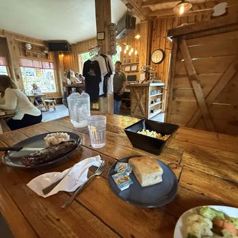 Showdowns Restaurant - Inside the restaurant - The image shows a wooden table inside a restaurant with rustic decor, including wooden walls and furniture. On the table are plates of food, a glass of water, a black bowl of popcorn, and a slice of bread with butter packets. In the background, diners are seated at tables, and a staff member stands near a counter with hanging shirts. The restaurant has large windows with red checkered curtains, and warm lighting illuminates the space. - Music Venues Near Me - Tropic, Utah