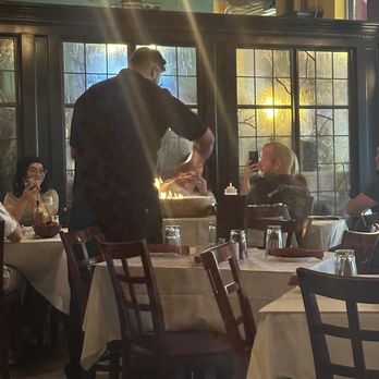 Cafe Bionda - A chef is standing at a restaurant table, preparing food in front of diners. Several guests are seated, some taking photos or videos with their phones, while others watch. The restaurant features large windows with a view of trees outside, and warm lighting fixtures hang from the ceiling. The tables are set with glasses, plates, and napkins, creating a casual dining atmosphere. - Italian Near Me - Chicago, Illinois