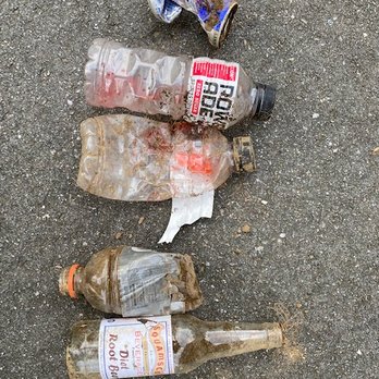 The Difference Landscapes - Trash thrown in my yard by employees; they tried to bury it. - Five empty bottles and a crushed soda can are scattered on a paved surface, some with labels partially visible and others dirty or damaged. - Landscaping Near Me - Lee, New Hampshire