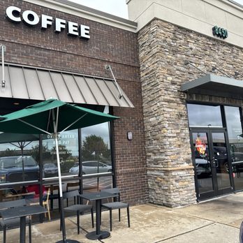 Starbucks - The photo depicts the front exterior of a Starbucks Coffee location, featuring a brick and stone facade. There are outdoor tables with chairs and green umbrellas set up on the sidewalk for outdoor seating. The storefront has large glass doors and windows, with signage indicating the business name. The parking lot and cars are visible through the windows, and the building's address is displayed on the corner. - Coffee & Tea Near Me - Irving, Texas