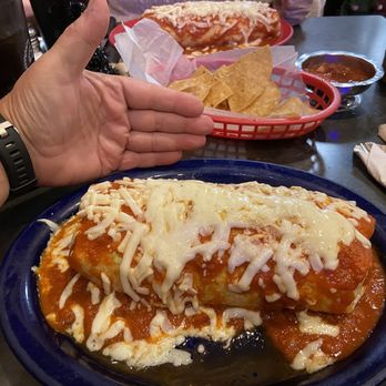 Tacos El Norte - Burrito Suizo Especial - A plate of cheese enchiladas topped with melted cheese and red sauce is placed on a dark table. A person's hand is visible near the plate, and in the background, there are tortilla chips, a small bowl of salsa, and another dish with cheese and sauce. The setting appears to be a Mexican restaurant. - Mexican Near Me - McHenry, Illinois