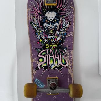 Maven - Staab deck. - A skateboard with a purple deck displays a graphic of a snarling, clown-like face with wild hair and exaggerated features. The deck has visible scratches and wear, with the graphic partially faded. The skateboard is mounted vertically on a white wall, with yellow wheels and metal trucks visible. The design includes vibrant colors and graffiti-style text, indicating a street or skateboarding culture theme. - Shoe Stores Near Me - Burlington, Vermont