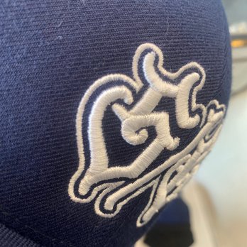 Arosha - The image shows a close-up of navy blue fabric with a white embroidered design. The embroidery appears to be a stylized or abstract pattern, with visible stitching detail. The fabric has a textured appearance, and the background is blurred, indicating the focus is on the embroidered area. - Graphic Design Near Me - Goleta, California