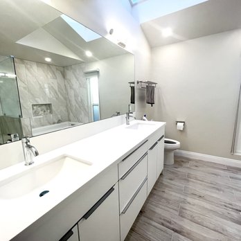 I Do Remodeling - Master Bathroom - The bathroom has a white double vanity with two integrated sinks and modern chrome faucets, a large mirror above the vanity, and a light-colored wood-look floor. A bathtub is visible in the background, along with a glass-enclosed shower area with marble tiles. Natural light enters through a window and a skylight, illuminating the space. The walls are painted white, and there are towel racks and a toilet nearby. - General Contractors Near Me - Fullerton, California