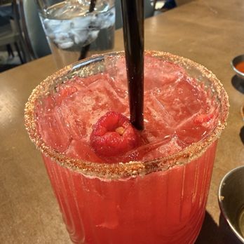 Hash Kitchen - Californication - A red cocktail with ice cubes and a cherry garnish is served in a glass with a salted rim. The drink has two black straws and is placed on a wooden table. In the background, there is a glass of water with ice and a straw, along with some small bowls. The setting appears to be a restaurant or bar. - New American Near Me - Peoria, Arizona