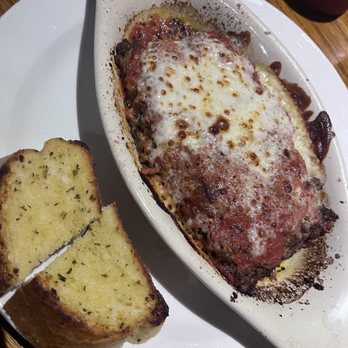 The Bay - Lasagne - A baked dish featuring melted cheese and tomato sauce, likely a lasagna or similar casserole, served in an oval ceramic dish. The dish has a browned, bubbly cheese topping and visible sauce around the edges. Two slices of garlic bread with herbs are placed on a white plate beside the dish. The setting appears to be a restaurant table with a wooden surface. - New American Near Me - Milwaukee, Wisconsin