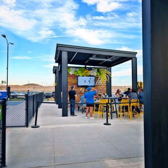 Serve - An outdoor seating area features a modern black metal shelter with a flat roof, decorated with balloons and a TV screen. Several people are seated at tables with yellow chairs, while others stand nearby. The area is enclosed by a black fence, with a parking lot and distant landscape visible beyond. The sky is bright blue with scattered white clouds, indicating clear weather. - Pickleball Near Me - El Paso, Texas