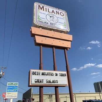 Milano Cafe - Sign - The image shows a tall, freestanding sign for Milano Italian Restaurant, featuring the restaurant's name, logo, and offerings such as steaks, pasta, seafood, and spirits. Beneath the main sign are two message boards, one with a message about mojitos and salads. The sign is set against a clear blue sky, with a parking lot and several vehicles visible at the base, along with other commercial signs nearby. - Italian Near Me - Lima, Ohio
