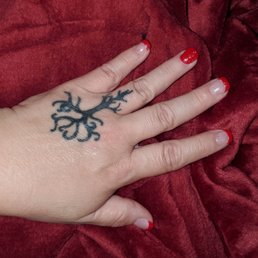 Left hand with red French tip on all but ring finger and a sparkling overlay. - A hand with a black tattoo of a tree on the back is shown with red French tip nails and clear glitter accents, resting on a red velvet fabric.