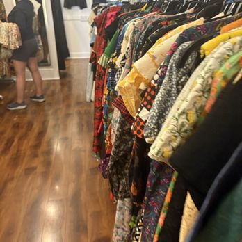 Vanishing Point - The image shows a clothing rack filled with a variety of colorful garments, including dresses and shirts, inside a boutique store. The store has wooden flooring, and a person is seen browsing or shopping in the background, near a mirror and other clothing displays. - Used, Vintage & Consignment Near Me - Oakland, California