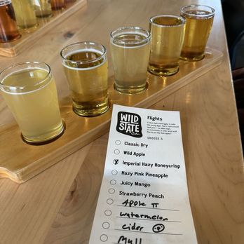 Wild State Cider - Flight - Five small glasses of cider are placed on a wooden tasting board at Wild State Cider, with a flight menu nearby listing various cider flavors. - Cideries Near Me - Duluth, Minnesota