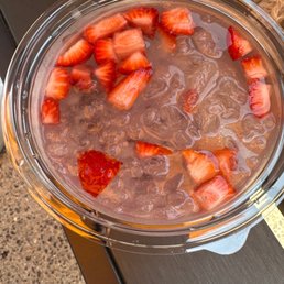 Doggie water bowl with strawberries and ice water. - A clear plastic cup holds iced water with ice cubes and sliced strawberries floating on the surface. The cup is positioned on a dark, textured surface, possibly a table or countertop. The strawberries are cut into small pieces and are partially submerged in the water. The image is taken from above, showing the contents clearly.