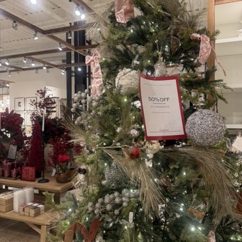 Pottery Barn - A Christmas tree is decorated with silver and glittery ornaments, pink ribbons, and string lights inside a store. The tree is surrounded by store displays with holiday-themed items such as flowers, candles, and gift boxes. A sale sign on the tree indicates a 50% discount on all ornaments. The store interior features a high ceiling with exposed beams and track lighting, creating a festive shopping environment. - Furniture Stores Near Me - Montvale, New Jersey