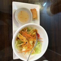 Part of main meal - A salad with lettuce, carrots, and dressing is served in a white bowl, accompanied by a spring roll and a small bowl of dipping sauce on a rectangular plate.
