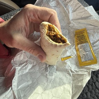 Wicked Brew - Inside of breakfast burrito - A hand grips a breakfast burrito that has been partially eaten, revealing a filling of meat and possibly other ingredients. The burrito is wrapped in a white paper with a honeycomb pattern, and a yellow sauce packet is placed on the paper. The setting appears to be inside a vehicle, with a textured gray surface and a seat visible in the background. - Coffee & Tea Near Me - Moab, Utah