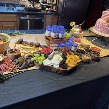 Tricky Foods - More of grazing table - The image shows a grazing table with a variety of fresh fruits, cheeses, nuts, and charcuterie arranged on a table covered with a black cloth and a beige runner. Yogurt cups are stacked in the center, and a pink decorated cake is placed on a stand in the background. The setting appears to be in a kitchen or event space with wooden cabinets and a stainless steel dishwasher visible behind the table. - Caterers Near Me - Madison, Wisconsin