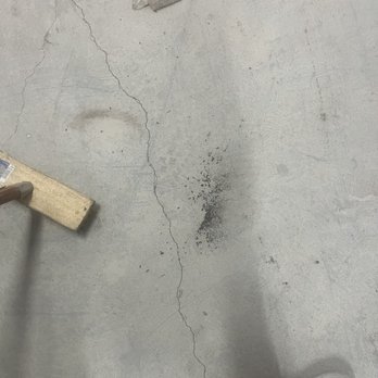 Extra Space Storage - Rat poop from the unit - Concrete floor with prominent cracks running across the surface, dark stains and spots, and small scattered debris such as a plastic bag and a piece of wood. The lower right corner shows part of a shoe, indicating a person standing nearby. The setting appears to be an industrial or storage area. - Self Storage Near Me - Bay Shore, New York