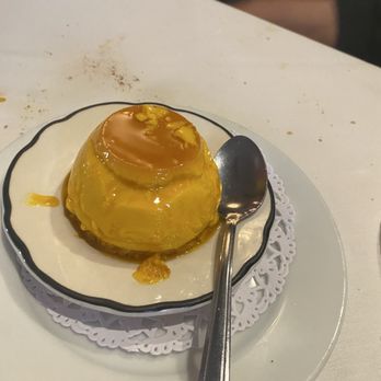 Casa Luis - Flan, yum - A caramel-topped flan dessert is served on a small decorative plate with a spoon, placed on a larger white plate with a doily. - Spanish Near Me - Smithtown, New York