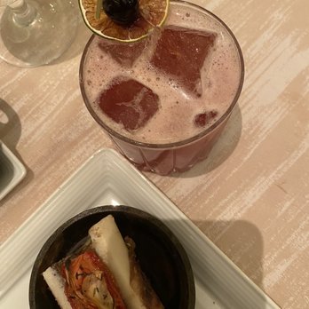 PY Steakhouse - Drinks - A pinkish cocktail with ice cubes and a garnish of dried citrus, cherry, and skewer is placed on a table. A small appetizer with cheese, tomato, and herbs is also visible. - Steakhouses Near Me - Tucson, Arizona