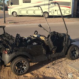 A severely burned golf cart is situated on a sandy patch next to a paved sidewalk, with a recreational vehicle and a building in the background.