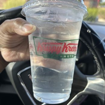 Krispy Kreme - Moldy cup of water - A transparent plastic cup with Krispy Kreme branding, containing water, is held by a person's hand inside a vehicle. The background shows part of the car's interior and a blurred outdoor scene with buildings and greenery. - Donuts Near Me - Manassas, Virginia