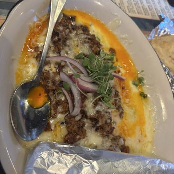 Sagrado - A Mexican dish served on a white oval plate, topped with ground meat, melted cheese, and garnished with red onion slices and microgreens. A metal spoon rests on the plate, and foil wrapping is visible at the bottom. The background includes a menu and other table items, indicating a restaurant setting. - Mexican Near Me - Waco, Texas
