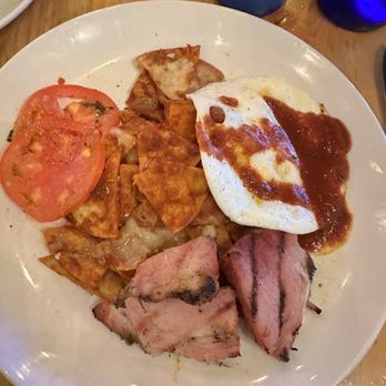 M'tucci's Italian - The chilaquiles! - A breakfast plate features fried eggs with sauce, sliced tomatoes, grilled meats, and potato chips, arranged on a white round plate. The plate is on a wooden table with drinks in the background, including a blue glass and a dark-colored glass. - Italian Near Me - Albuquerque, New Mexico