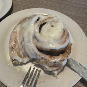 Henhouse Cafe - Cinnamon Roll freshly baked - A cinnamon roll topped with white icing is placed on a white plate at a restaurant table, with a fork and a knife nearby. - Cafes Near Me - Gilbert, Arizona
