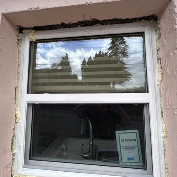 Flawless Results - The image shows a double-pane window with a white frame installed in a pink exterior wall. The wall has visible foam insulation around the window frame, indicating recent installation or repair work. The window reflects the sky, clouds, and trees outside, and a sticker from ProVia is visible on the lower pane. The surrounding wall surface appears textured and unfinished in some areas. - Windows Installation Near Me - Albuquerque, New Mexico
