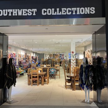 Southwest Collections - Entrance - The image shows the storefront of Southwest Collections, a retail store specializing in southwestern products. The store's entrance is framed by mannequins dressed in southwestern-style clothing and accessories. Inside, the store displays a variety of furniture, jewelry, and decorative items reflecting Native American designs. The interior is well-lit with ceiling spotlights, and customers are browsing the merchandise. The store appears spacious and organized, with items arranged on walls and tables. - Furniture Stores Near Me - Albuquerque, New Mexico