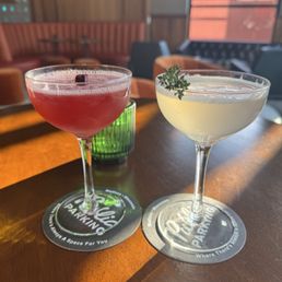Two cocktails are placed on a wooden table inside a bar with warm lighting, wooden wall decor, and large windows.