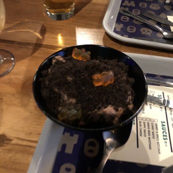 The Beer Keep - Pudding - A dessert served in a black bowl, topped with crushed cookies and gummy candies, is placed on a tray on a wooden table. The setting includes a glass of beer, a tray with utensils, and a menu or paper with drink options. The environment appears to be a casual dining or bar area. - Beer Bar Near Me - Buffalo, New York