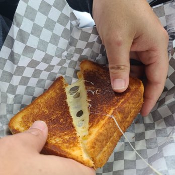 Chill...The Milkshake Bar - The Texas grilled cheese - A person is holding a grilled cheese sandwich with melted cheese stretching between the two halves. The sandwich is on checkered paper, and the person's hands are visible, with one hand pulling the sandwich apart. The background suggests a casual dining setting. - Ice Cream & Frozen Yogurt Near Me - Spring, Texas