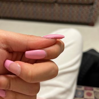 VIP Nails - And they are already chipping. (They are gel too). - The image features a close-up of a hand with well-maintained, pink, glossy nails. The background includes a patterned sofa with multicolored upholstery and a person wearing white pants, sitting in an indoor setting. The focus is on the nails and hand, with the background slightly blurred. - Nail Salons Near Me - Amherst, Massachusetts