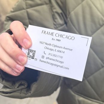 Frame Chicago - A person in a gray quilted jacket holds a business card for Frame Chicago, displaying contact details and a QR code. - Framing Near Me - Chicago, Illinois