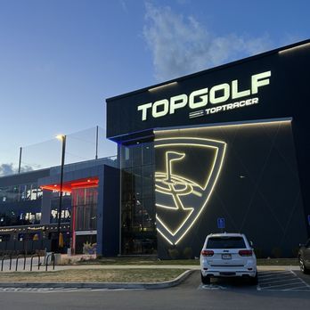 Topgolf - The photo captures the exterior of Topgolf in Canton, MA, during dusk, with a clear sky overhead. The building has prominent illuminated signage displaying 'TOPGOLF' and a large, illuminated logo of a golf tee and ball. The parking lot in front includes a white SUV and a few other vehicles, with a grassy area and bike racks nearby. The modern architecture features glass and dark panels, with some interior lighting visible through the windows. - Golf Near Me - Canton, Massachusetts