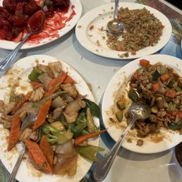 Family meal - Four plates of Chinese takeout dishes are arranged on a table, each containing different stir-fried vegetables, rice, and meat. The dishes include mixed vegetables with chicken, fried rice, and sweet and sour chicken with sauce. Spoons are placed on each plate for serving. The table surface is light-colored, and part of a decorative placemat or menu is visible underneath.