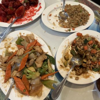 Friendly House - Family meal - Four plates of Chinese takeout dishes are arranged on a table, each containing different stir-fried vegetables, rice, and meat. The dishes include mixed vegetables with chicken, fried rice, and sweet and sour chicken with sauce. Spoons are placed on each plate for serving. The table surface is light-colored, and part of a decorative placemat or menu is visible underneath. - Chinese Near Me - Turlock, California