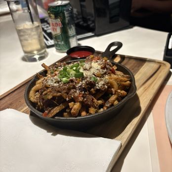 Nosso - Oxtail loaded fries - A cast iron skillet filled with loaded fries, garnished with chopped green onions and grated cheese, served on a wooden board at a restaurant. - Cocktail Bars Near Me - Oakland, California
