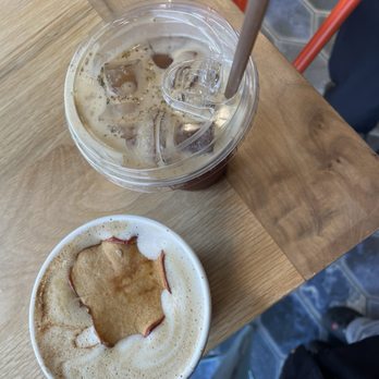 Cloud Walking Coffee - Apple Chai Latte (w/ shot of espresso) and Solstice Tonic - The image shows two beverages on a wooden table in a cafe. The first is an iced coffee with ice cubes, a straw, and a clear plastic lid. The second is a hot coffee with foam and a cookie placed on top of the foam. The background includes a tiled floor and part of a person’s leg, indicating a casual cafe environment. - Chocolatiers & Shops Near Me - South Bend, Indiana