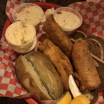 Ron's Place - Fish and baked potato with buttah, cole slaw and tartar - A red basket lined with checkered paper contains fried fish fillets, a baked potato with a split open revealing its interior, coleslaw, tartar sauce, and slices of dark bread. Two lemon wedges are placed at the front of the basket. The setting appears to be a casual dining environment, possibly at a restaurant or bar. - Pubs Near Me - Kenosha, Wisconsin
