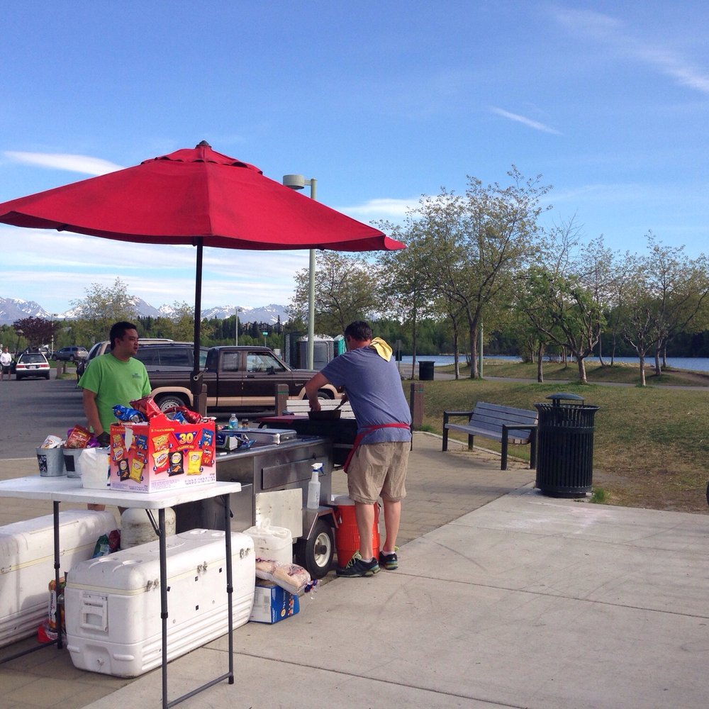 Denali Sausage - two men standing at a table with a red umbrella - Food Stands Near Me - Anchorage, Alaska Denali Sausage - two men standing at a table with a red umbrella - Food Stands Near Me - Anchorage, Alaska