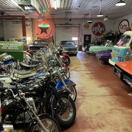 Dinner and Museum - The image shows a spacious indoor area with a collection of vintage motorcycles in the foreground, featuring windshields and chrome details. Behind the motorcycles, there are classic cars, including a black car and a light green car, parked along the wall. The space is decorated with vintage signs, such as a Texaco neon sign and Derby signage, creating a nostalgic atmosphere. The ceiling has metallic panels with hanging lights, and the floor appears to be a worn, reddish-brown surface.